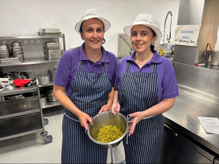 Two people wearing aprons in a commercial kitchen holding a bowl of food