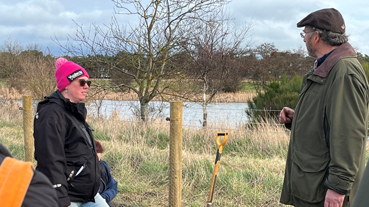 Two people stand chatting next to a fenced off pond. 