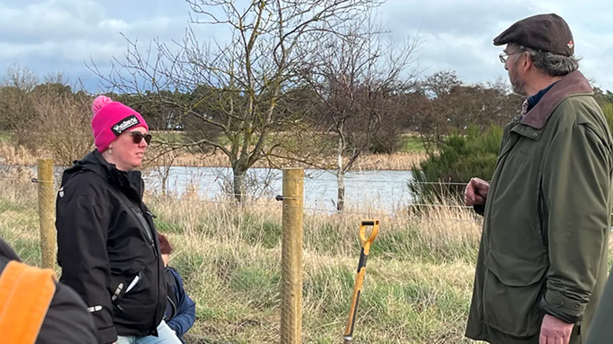 Two people stand chatting next to a fenced off pond. 