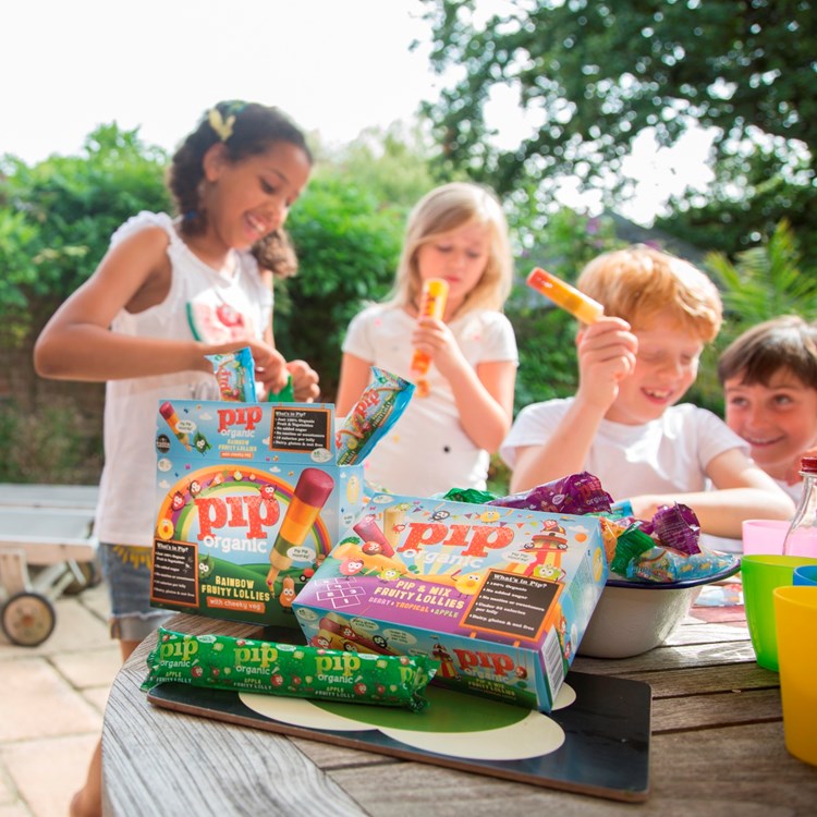 Kids eating Pip Organic ice lollies in the background, with a garden table laden with Pip Organic products in the foreground