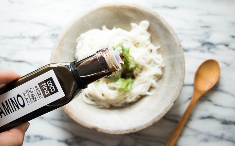 Someone holding a bottle of Cocofina amino oil and drizzling it on a bowl of noodles with a wooden spoon next to it set on a marble countertop