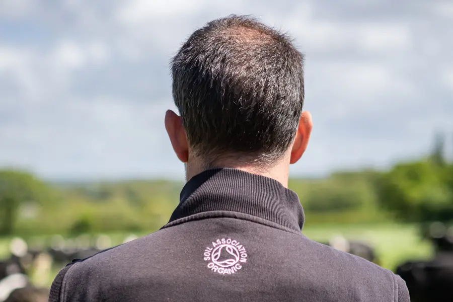 A man wearing a jumper emblazoned with the Soil Association organic symbol looks out over a field of cows