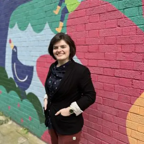 A smiling woman with short hair wearing a blazer and standing in front of a colourful mural on an exterior wall