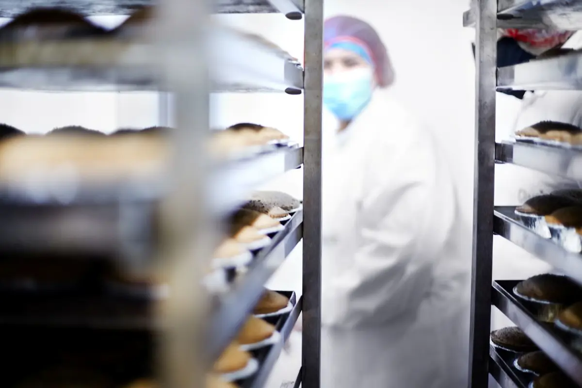 Person wearing a protective white coat, face mask, and hair net in a factory environment, near trays of pies