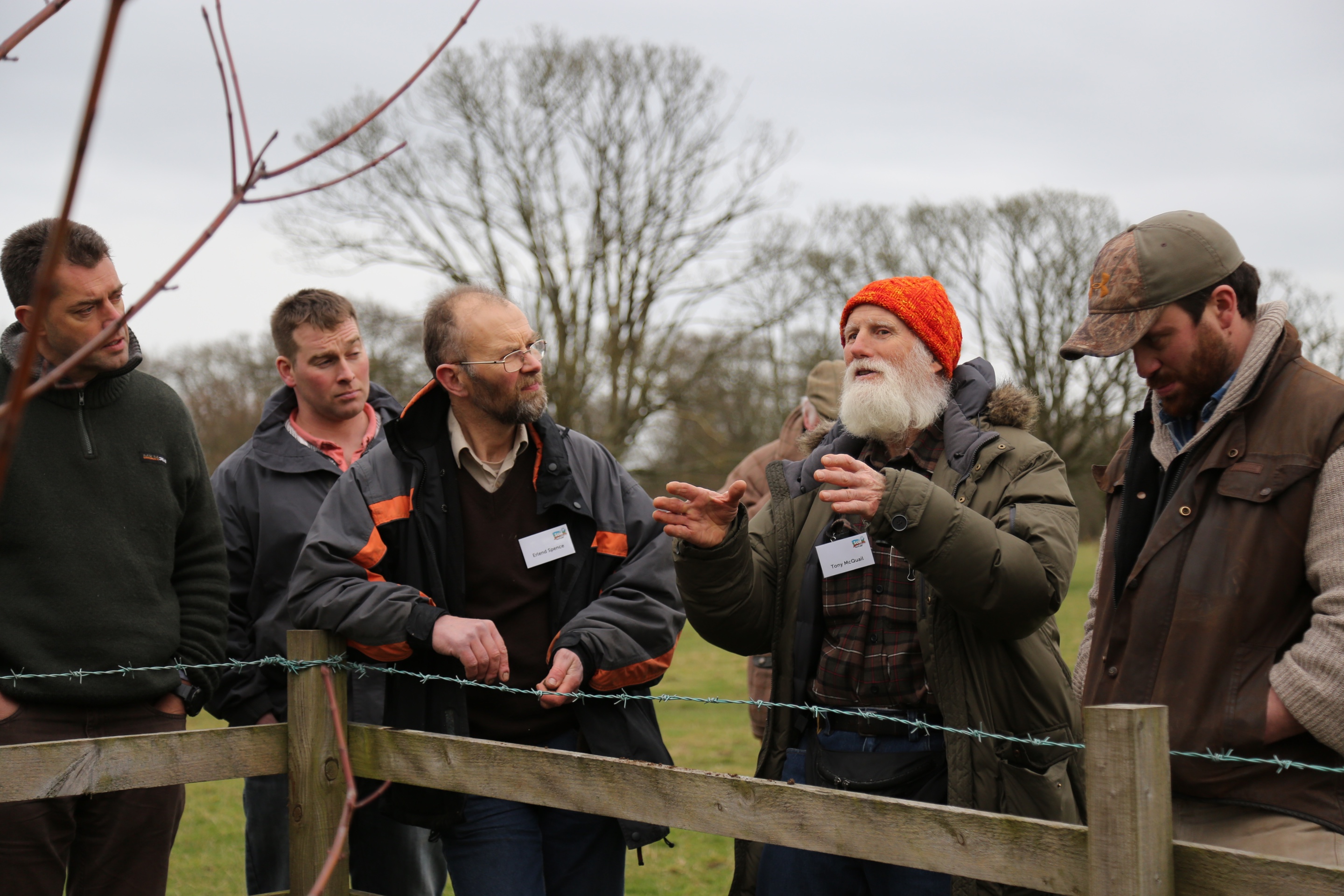 Five men in warm outdoor clothing stand at a fence. One is talking animatedly while the others listen. 