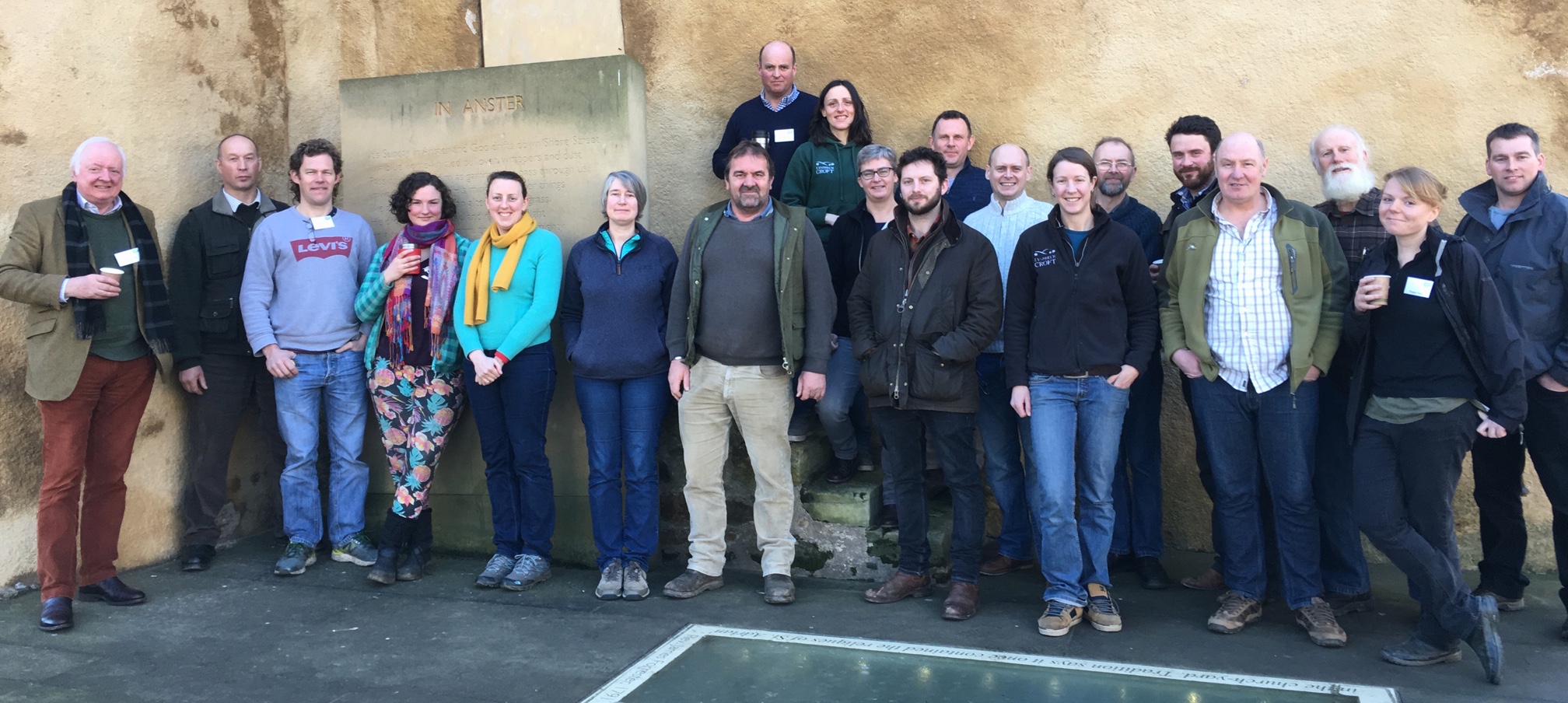 Twenty people stand smiling at the camera, against a sandy coloured concrete wall. 