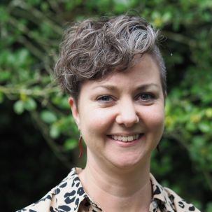 A smiling woman with short, curly hair and a leopard print shirt standing near trees