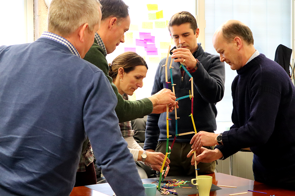 A group of 5 people gathered around a table participate in a tower building activity