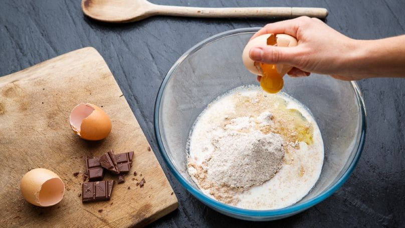 Person cracking an egg into a bowl of flour, with a wooden spoon, used eggshells, and chunks of chocolate nearby