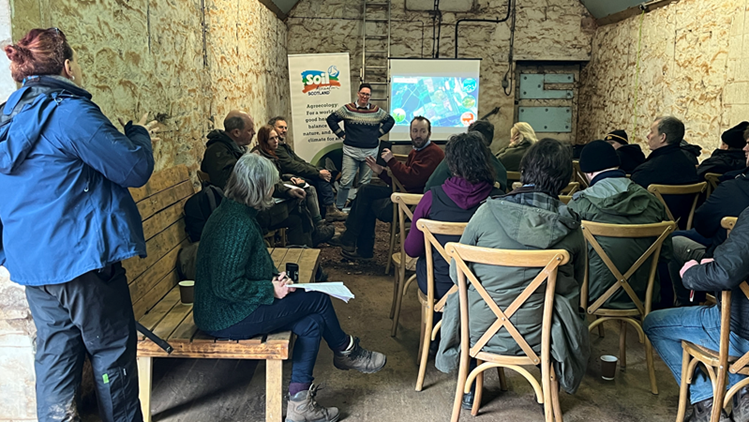 A group of around 15 people attend a presentation in a room with chipboard walls. A Soil Association banner stands at the front. 