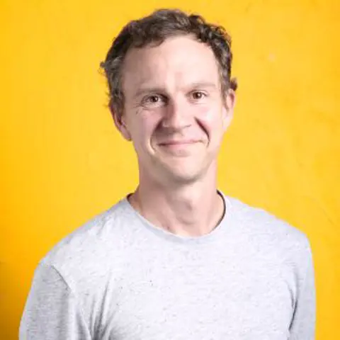 A smiling man with short brown hair and brown eyes smiling against a bright yellow background