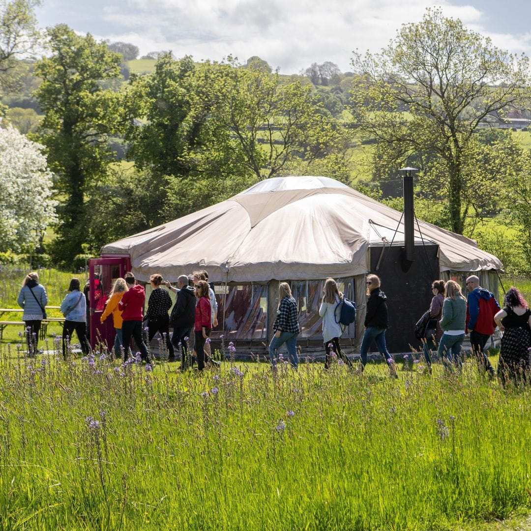 A yurt in a field, with a large gathering of people heading towards it