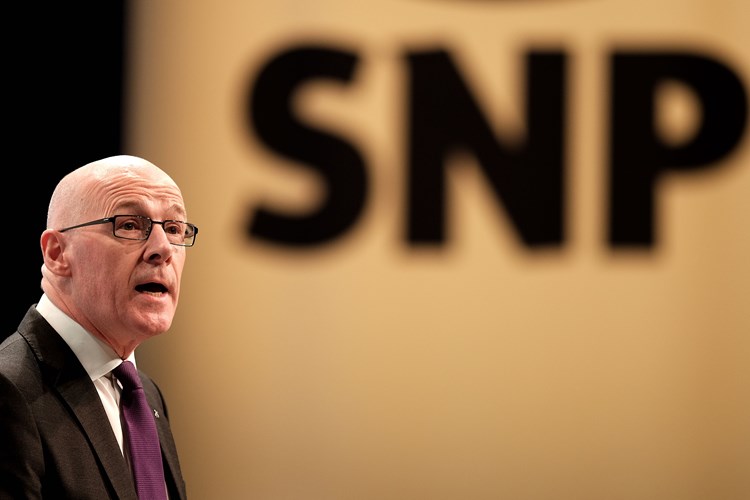 John Swinney speaks in front of a large, out of focus SNP logo in background. 