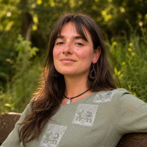 A woman with long brown hair sits on an outdoor bench surrounded by nature
