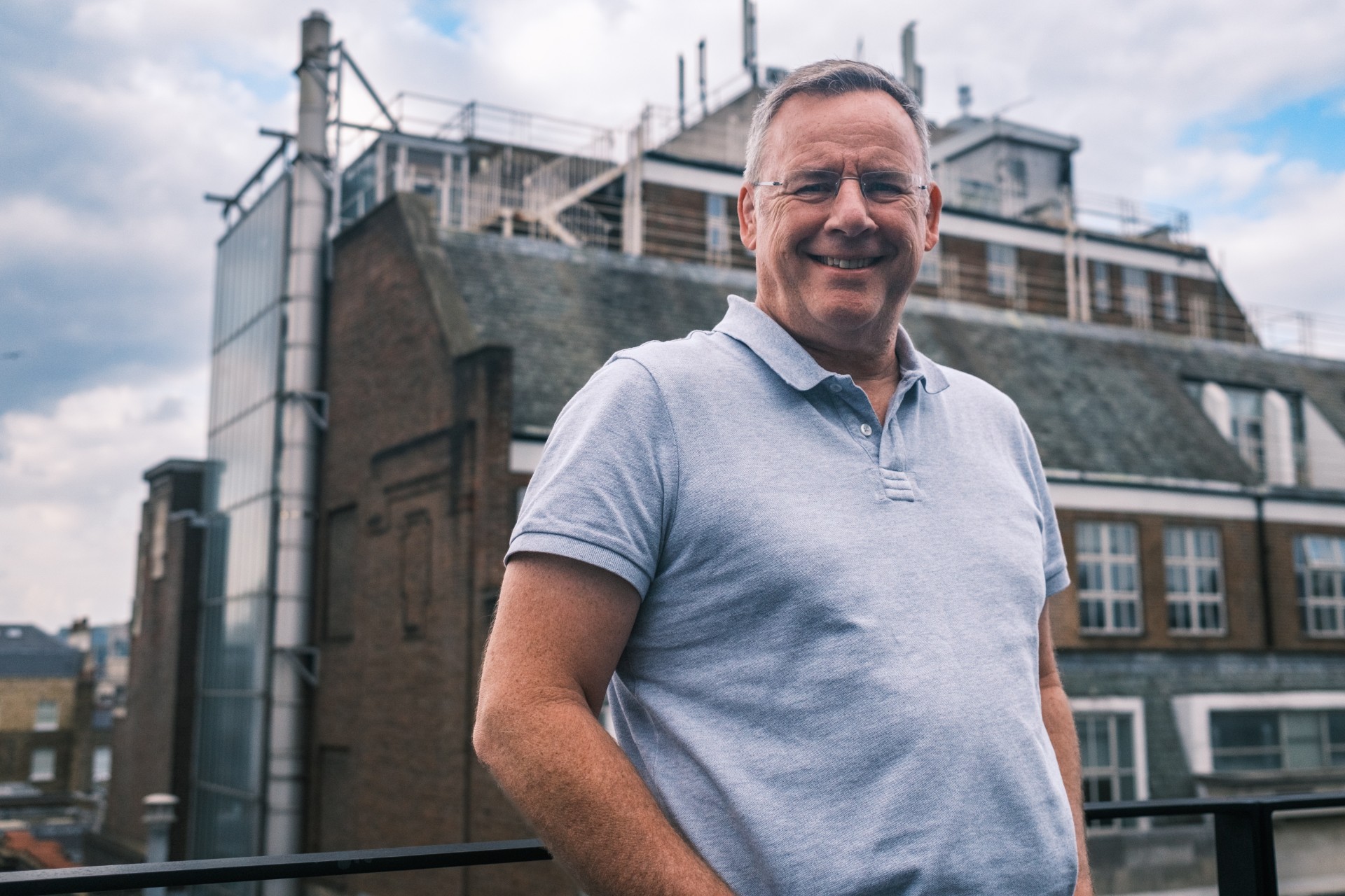 A man standing on a rooftop, wearing glasses and a grey polo shirt