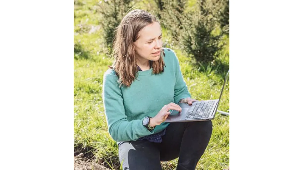 Ichsani Wheeler, OpenGeoHub Foundation kneeling in a field with one hand using the touchpad of a laptop