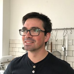 A smiling man standing in a kitchen with black hair and black framed glasses