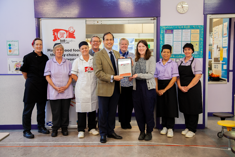 A man and woman smile at the camera while holding a framed certificate, surrounded by colleagues wearing kitchen overalls and aprons.  